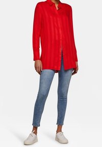 Camicia a maniche lunghe rossa con bottoni e righe verticali, abbinata a jeans skinny di un blu chiaro e sneakers bianche. Tessuto liscio, design con colletto.