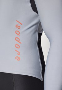 Sivé cyklistické dlhé tričko s čiernym bočným panelom, s vertikálnym oranžovým logom "Isadore". Hladká, elastická textúra látky.