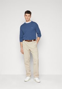 Blauwe gebreide trui met geribbelde textuur, crèmekleurige chinos, een bruine riem en witte sneakers. Model poseert tegen een effen lichte achtergrond.
