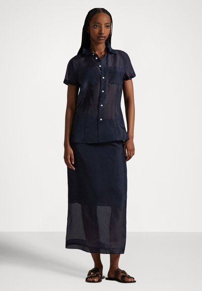 Sieviete, tērpusies tumši zilā caurspīdīgā īsroku krekla ar pogām un atbilstošā caurspīdīgā potīšu garuma svārkā, valkājot brūnas sandales, stāv pret vienkāršu fonu.