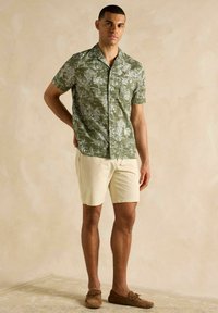 Zelená krátkopyjámková košeľa s potlačou palmy a plachetnice, spárovaná s béžovými šortkami a hnedými sandálmi. Hladká textúra.