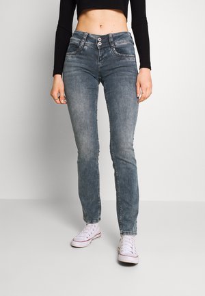 Femme portant un jean bleu délavé taille haute avec fermeture à double bouton, un crop top noir à manches longues et des baskets blanches, debout sur un sol clair.