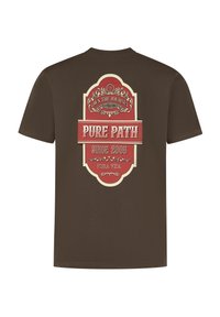 Bruine t-shirt met een groot grafisch ontwerp in rood en crème, met de tekst "BEHEERS DE REIS" en "PURE PAD, SINDS 2008" in decoratieve lettertype.