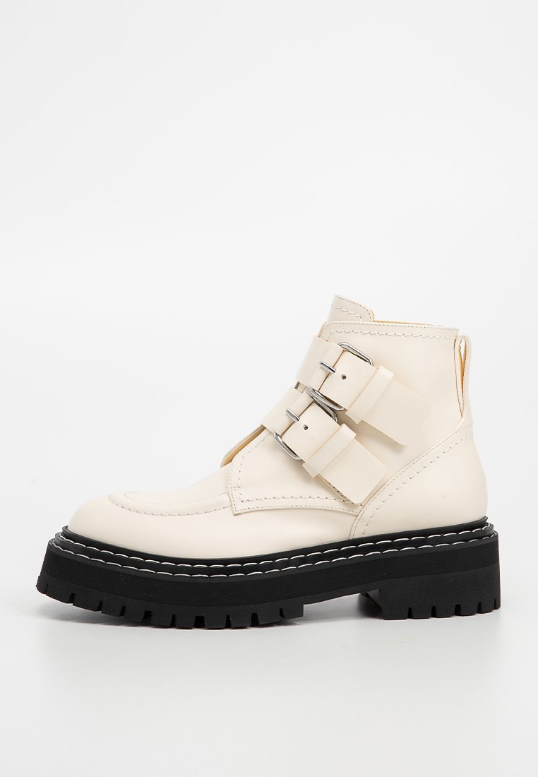 Kremowe skórzane botki za kostkę z czarną grubą podeszwą i podwójnymi srebrnymi klamrami po bokach, zaprezentowane na białym tle.