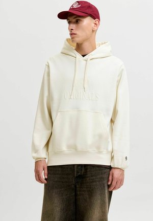 Jeune homme portant un sweat à capuche crème avec le texte "ORIGINALS" et une casquette de baseball bordeaux, debout devant un fond clair uni.