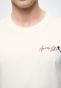 Camiseta de algodón color crema con cuello redondo, que presenta el texto bordado en rojo y azul "Après Ski" y un diseño de bastón de esquí en el pecho.