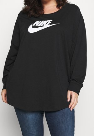Femme portant un haut Nike noir à manches longues avec logo blanc, associée à un jean bleu, debout devant un fond uni.