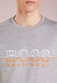 Šedá mikina s výrazným vyšitým textem v oranžové a bílé barvě, který zní "BOSS HUGO BOSS." Látka je měkká a texturovaná s designem kulatého výstřihu.