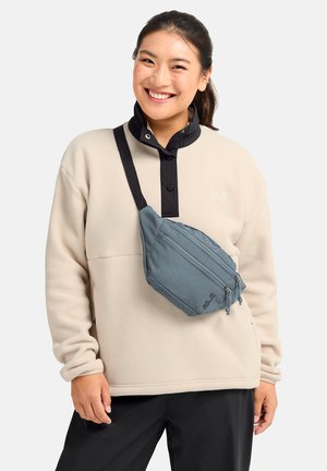 Junge Frau lächelnd, trägt eine beige Fleecejacke, schwarze Hose und eine blaue Umhängetasche mit zwei Reißverschlussfächern.