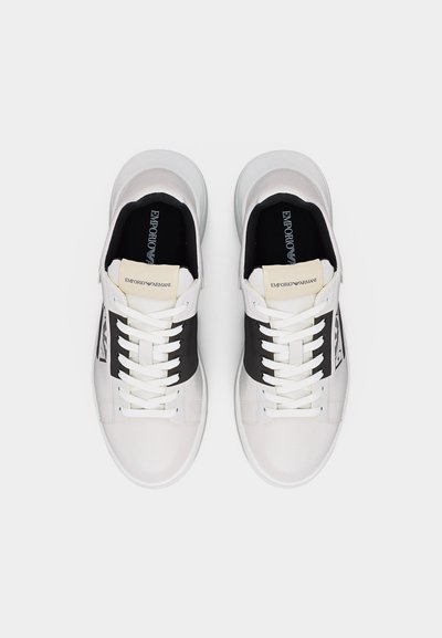 Ζευγάρι λευκά αθλητικά παπούτσια με μαύρα πλαϊνά πάνελ, λευκά κορδόνια και μπεζ ετικέτες στη γλώσσα με την επωνυμία "Emporio Armani", ορατά από πάνω.
