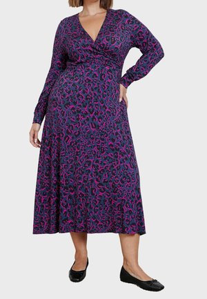 Vrouw draagt een midi-jurk met lange mouwen, paarse en roze luipaardprint, met een overslagstijl lijfje en zwarte ballerina's.
