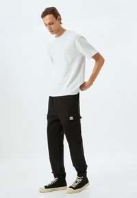 Wit, kortmouwshirt met een rauwe zoom, gecombineerd met zwarte cargobroek met zijzakken en een labelpatch, gedragen met zwarte sneakers.