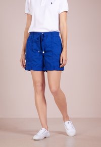 Kvinna iklädd blå dragsko-short, vit polo och vita sneakers som står framför en neutral bakgrund.