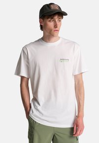 Λευκό βαμβακερό T-shirt με στρογγυλή λαιμόκοψη και κοντά μανίκια· διαθέτει πράσινο και μαύρο λογότυπο στην αριστερή πλευρά του στήθους.