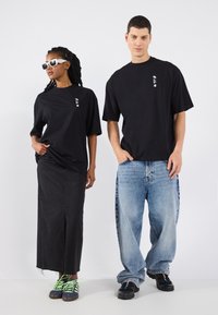 T-shirts noirs surdimensionnés avec des détails graphiques blancs, associés à une jupe en denim noire et un jean baggy bleu clair. Les sneakers sont dotées de lacets verts fluo.