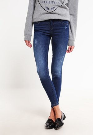 Femme portant un jean skinny bleu foncé avec des légères décolorations et des mocassins noirs brillants, assorti à un sweat-shirt gris.