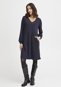 Navyblaues Kleid mit gelbem Karomuster, V-Ausschnitt, langen Ärmeln und seitlichen Taschen, kombiniert mit knielangen schwarzen Lederstiefeln.