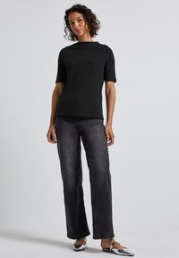 Kvinde iført en sort ribbet kortærmet top, falmede sorte brede jeans og sølvfarvede flade sko, stående foran en ensfarvet lys baggrund.