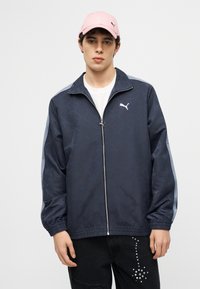 Νεαρός  άνδρας που φοράει ένα μπλε σκούρο ζακέτα Puma με ανοιχτό μπλε ρίγες, μαύρο παντελόνι με λευκά καρφιά και ένα ροζ καπέλο Puma, στέκεται μπροστά σε ένα απλό φόντο.