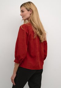 Blouse rouge à manches bouffantes en tissu texturé, avec un col rond et des poignets boutonnés, portée avec un pantalon noir.