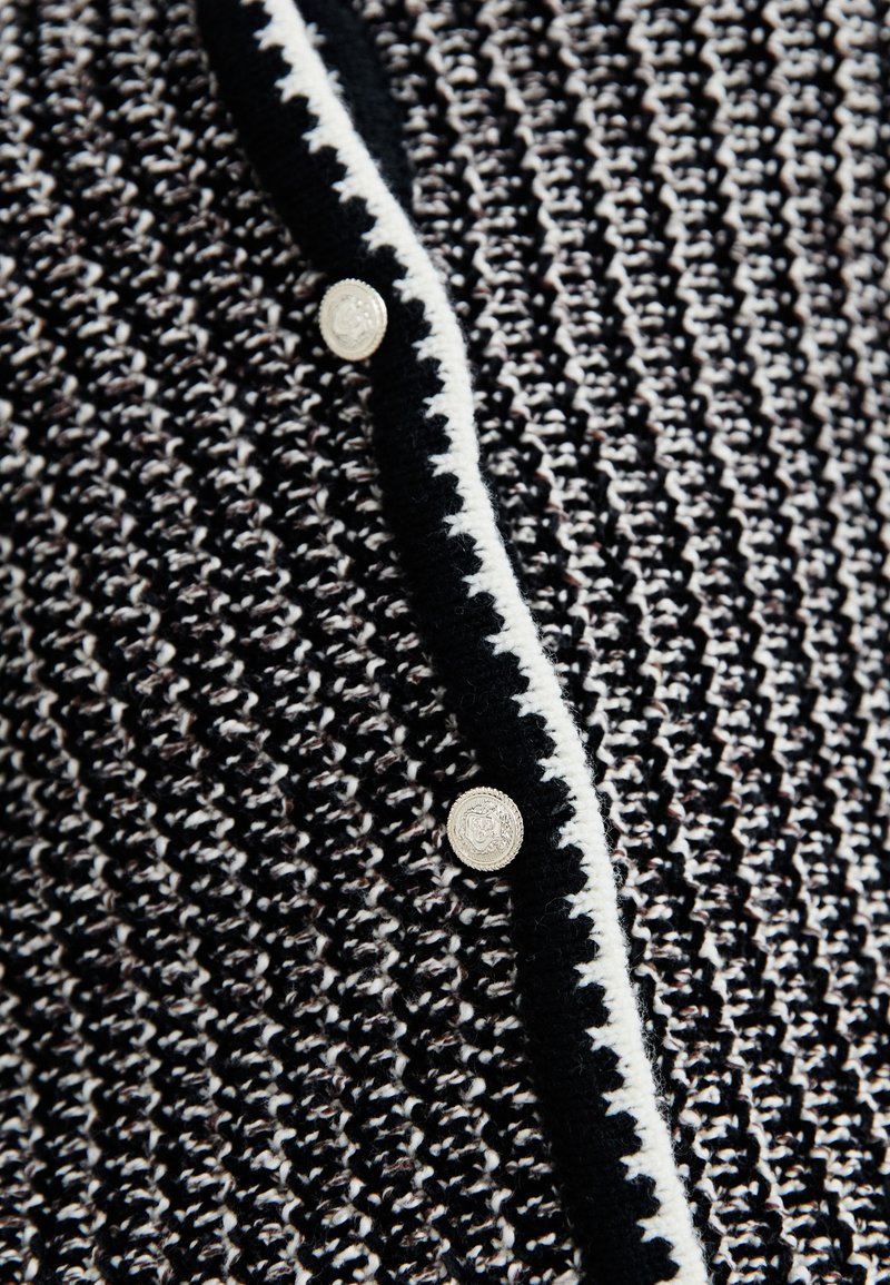 Schwarzer und weißer gemusterter Cardigan mit strukturiertem Strick, kontrastierenem Saum und silbernen Knöpfen, die die Vorderseite schließen.