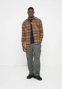 Camisa de franela a cuadros en color naranja y gris con dos bolsillos, combinada con pantalones cargo grises que tienen múltiples bolsillos y zapatillas negras.