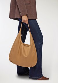 Borsa hobo in nylon color beige con una texture liscia, dotata di una tracolla ampia e di una chiusura con zip, portata da una persona che indossa jeans blu scuro e un blazer marrone.