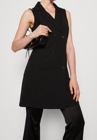 Schwarzes ärmelloses Blazer-Kleid mit doppelt geknöpftem Design, großen schwarzen Knöpfen und tailliertem Schnitt, kombiniert mit glänzenden schwarzen Hosen.