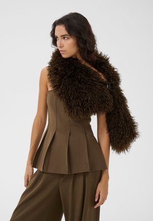 Vrouw draagt een mouwloze bruine geplooide top en bijpassende wijde broek met een grote donkerbruine pluizige bontsjaal over één schouder gedrapeerd.