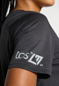 T-shirt de sport noir à manches courtes, doté d'un tissu texturé et d'un logo gris sur la manche. Le logo comprend "tcs", un emblème et le numéro "25".