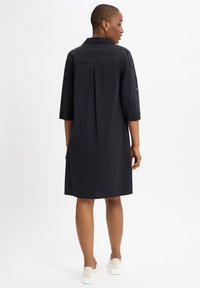 Zwarte jurk met een kraag en driedelige mouwen, met een plooien aan de achterkant, gemaakt van een gladde stof, gecombineerd met witte sneakers.