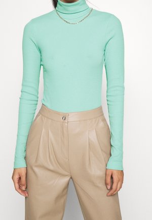 Femme portant un pull col roulé côtelé vert clair et un pantalon en cuir beige plissé taille haute, accessoirisé avec deux colliers en or.