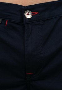 I pantaloni di cotone blu scuro presentano un bottone metallico rotondo con logo, cuciture di accento rosse e una texture liscia.