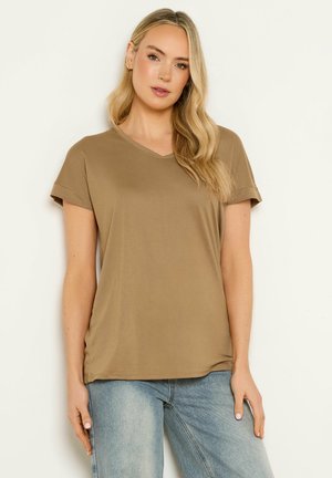 Femme aux longs cheveux blonds portant un t-shirt marron à manches courtes et col en V, et un jean bleu clair, debout devant un fond clair.