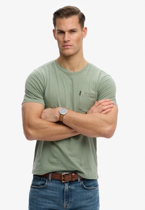 Mężczyzna o krótkich włosach, ubrany w zielony t-shirt z kieszenią, niebieskie dżinsy, brązowy pasek i zegarek na rękę, stoi ze skrzyżowanymi ramionami.