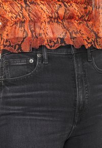 Top transparente em camadas com padrão de cobra em laranja e preto, combinado com jeans escuros de corte justo, apresentando um bolso frontal e detalhes em botão.
