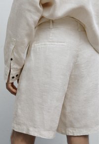 Pantaloni corti in lino beige con una texture liscia, dotati di una tasca posteriore e una vita aderente. L'orlo è dritto e leggermente sopra il ginocchio.
