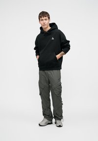 Sweatshirt preto com capuz, bolso frontal e logotipo, combinado com calças cargo cinza com múltiplos bolsos e um corte descontraído. Ténis cinza.