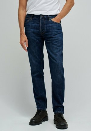 Hombre con vaqueros azul oscuro, camiseta blanca y botas negras con cordones, de pie con una mano en el bolsillo contra un fondo liso.