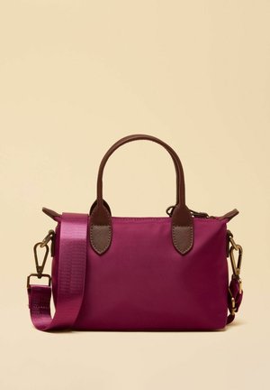 Geantă mică dreptunghiulară de culoare mov, cu mânere maro și curea lată de umăr detașabilă, pe un fundal bej.