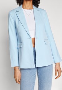 Světle modrý blazer se strukturovaným designem, klopami, zapínáním na jeden knoflík a předními kapsami. Nosený přes bílý zkrácený top.