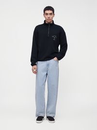 Mannequin homme portant un sweat-shirt noir à fermeture éclair partielle avec un petit logo et un jean large bleu clair avec des baskets noires, debout devant un fond uni.