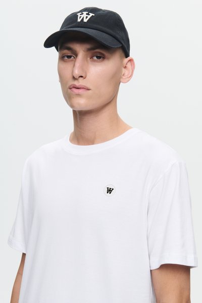 Jonge man met een zwarte pet en een wit T-shirt met een klein zwart "W"-logo op de voorkant, tegen een eenvoudige lichte achtergrond.