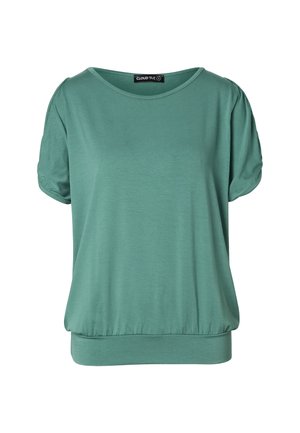 Femme blonde portant un haut bleu sarcelle à manches courtes avec des épaules découpées et un pantalon noir, debout devant un fond clair uni.