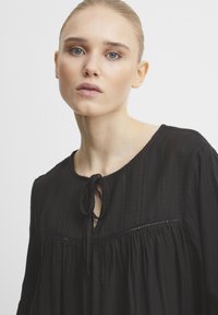 Blusa nera realizzata in tessuto leggero, con scollatura rotonda e fiocco frontale, righe verticali e dettagli arricciati.