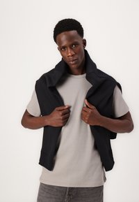 Grå bomulds t-shirt iført under en sort sweatshirt, der hænger over skuldrene; stoffet ser blødt ud med en afslappet pasform og minimalistisk design.