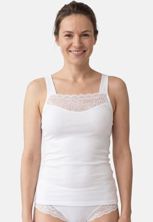 Lächelnde Frau, die ein weißes ärmelloses Oberteil mit Spitzendetail und passende Unterwäsche mit Spitzenbesatz trägt, vor einem einfarbigen Hintergrund steht.