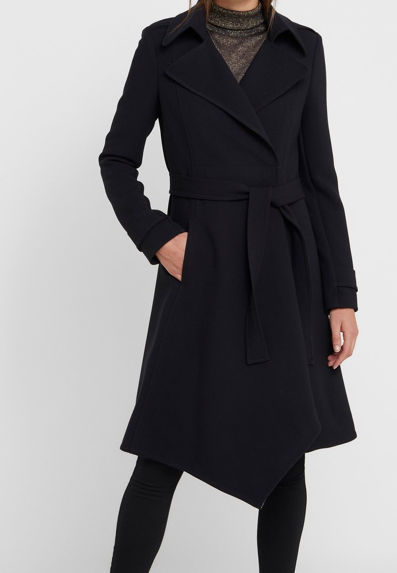 Mustassa trenssitakissa on tupla-kallellaan oleva muotoilu, vyösolki vyötäröllä ja levenevä helma. Se on valmistettu paksusta kankaasta ja siinä on suuret kaulukset ja sivutaskut.