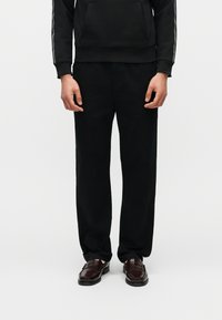 Άτομο που φοράει μαύρο φαρδύ παντελόνι, μαύρη κουκούλα με κορδέλα λογότυπου στα μανίκια και σκούρα καφέ δερμάτινα loafers, στέκεται σε λευκό φόντο.