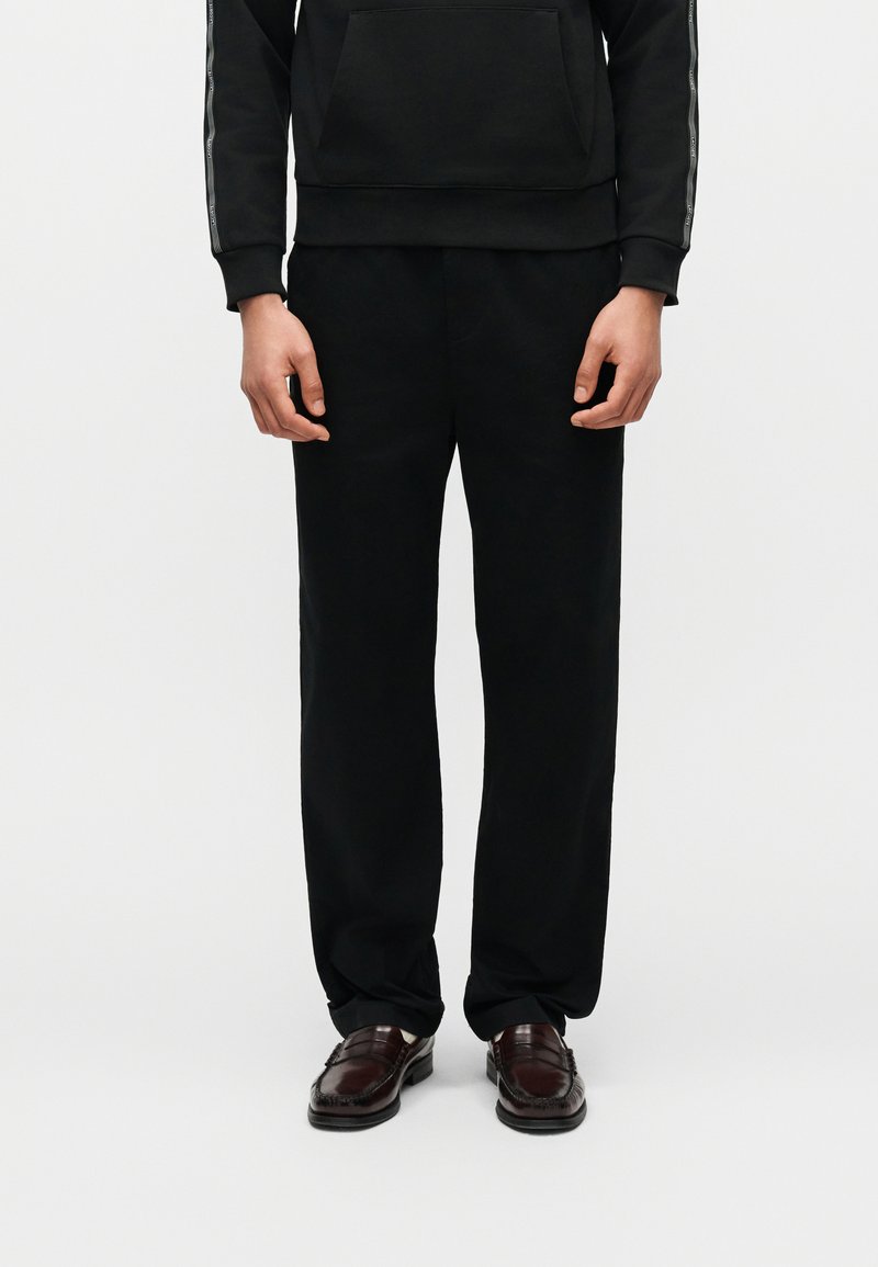 Άτομο που φοράει μαύρο φαρδύ παντελόνι, μαύρη κουκούλα με κορδέλα λογότυπου στα μανίκια και σκούρα καφέ δερμάτινα loafers, στέκεται σε λευκό φόντο.